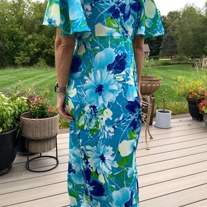 Puede incluir: Un vestido largo hasta el suelo con un vibrante estampado floral en tonos azul brillante, azul marino y blanco con toques de verde. La prenda tiene mangas cortas acampanadas y un corpi&ntilde;o ajustado que cae en una falda larga y recta.