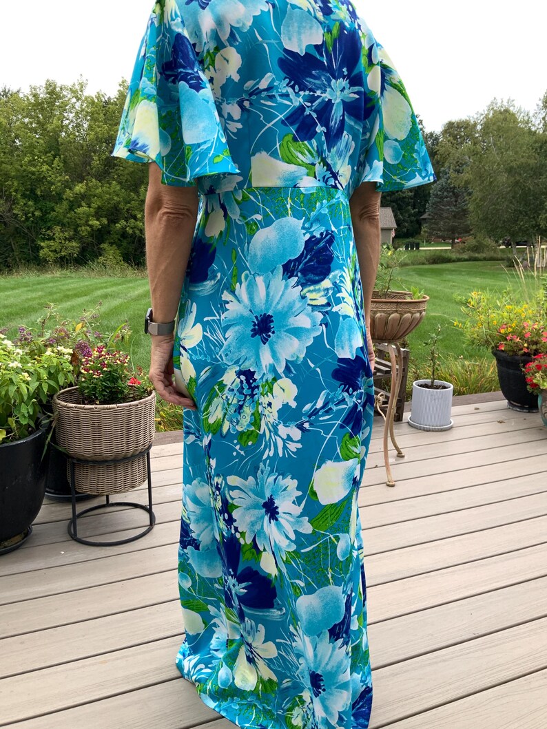 Puede incluir: Un vestido largo hasta el suelo con un vibrante estampado floral en tonos azul brillante, azul marino y blanco con toques de verde. La prenda tiene mangas cortas acampanadas y un corpi&ntilde;o ajustado que cae en una falda larga y recta.