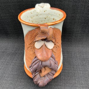 May include: Handmade ceramic mug with a whimsical face design. The mug features a white interior with brown speckles, an orange rim, and a sculpted face with a brown beard, nose, and eyebrows. The handle is white.