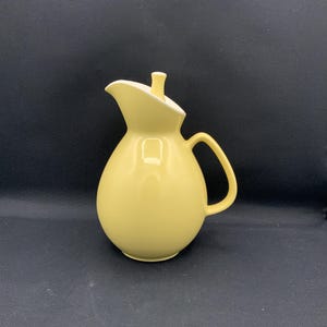 May include: A pale yellow ceramic pitcher with a rounded body, a curved handle, and a matching lid featuring a small knob handle. The pitcher has a wide, pointed pouring spout and a smooth, glossy finish, presenting a clean and simple design.