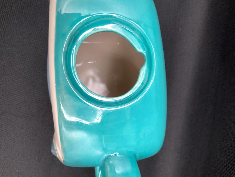 May include: A turquoise ceramic watering can with a large circular opening and a small handle. The watering can has a glossy finish and a light beige accent on the side. The interior is a light brown color.