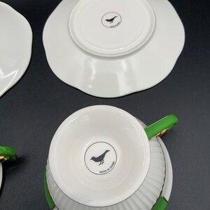 May include: A set of two white teacups with green and gold trim. The cups have a fluted design and a black bird logo on the bottom that reads "Made in China".