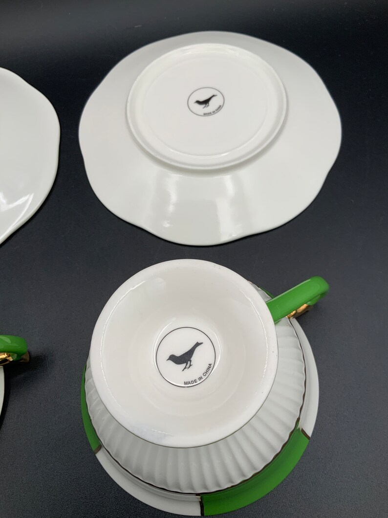 May include: A set of two white teacups with green and gold trim. The cups have a fluted design and a black bird logo on the bottom that reads "Made in China".
