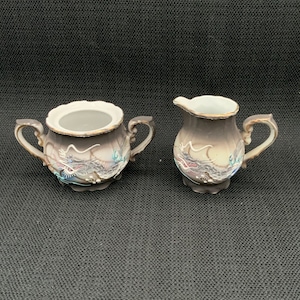 May include: A vintage porcelain creamer and sugar set with a dragon design. The creamer and sugar bowl are both decorated with a brown, gray, and blue dragon design. The set has a gold trim.