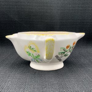 Puede incluir: Cuenco de hierbas de cerámica blanca con asa y pie pequeño. El cuenco está decorado con diseños florales amarillos, verdes y naranjas y la palabra "OREGANO". El cuenco tiene un borde festoneado.