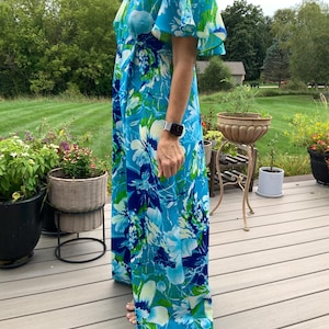 Puede incluir: Un vestido cruzado largo hasta el suelo con un estampado floral vibrante en tonos azul brillante, azul marino y blanco con toques de verde. La prenda tiene un escote en V y mangas cortas y sueltas que caen sobre los hombros y brazos.