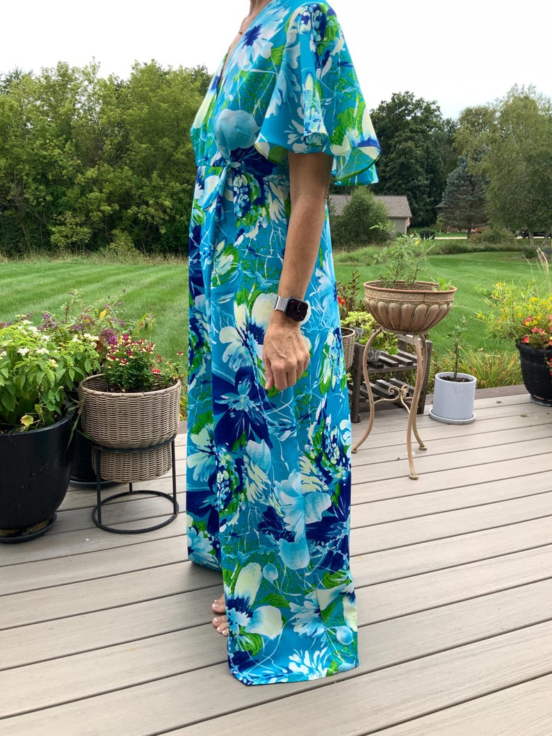Puede incluir: Un vestido cruzado largo hasta el suelo con un estampado floral vibrante en tonos azul brillante, azul marino y blanco con toques de verde. La prenda tiene un escote en V y mangas cortas y sueltas que caen sobre los hombros y brazos.