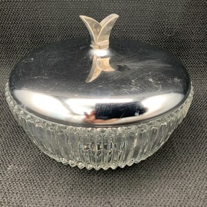 May include: A clear glass candy dish with a silver-colored lid and a decorative handle. The dish has a ribbed design and a scalloped edge. The lid is shiny and reflects the surrounding environment.