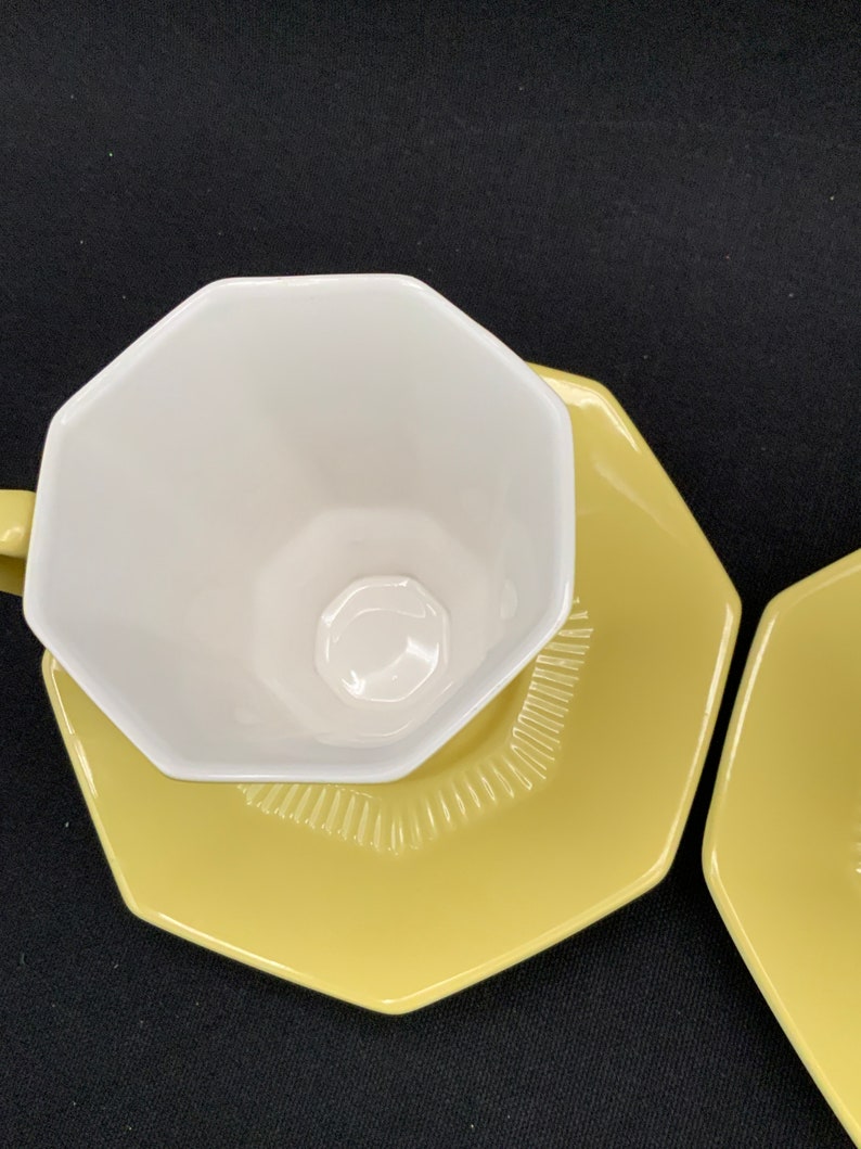 K&ouml;nnte beinhalten: Ein gelber achteckiger Unterteller mit einer wei&szlig;en achteckigen Teetasse darauf. Der Unterteller hat ein dekoratives Muster am Rand.