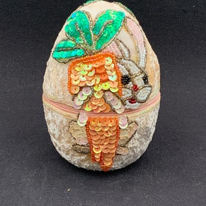 May include: A white egg-shaped trinket box with a velvet exterior. The box is decorated with sequins and beads in the shape of a rabbit and a carrot. The rabbit is white with pink ears and black eyes. The carrot is orange and green.