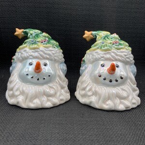 May include: Two ceramic snowman figurines. Each snowman has a white beard, a carrot nose, and black dot eyes. They wear green hats with gold star accents and are decorated with holly. The snowmen are on a dark background.