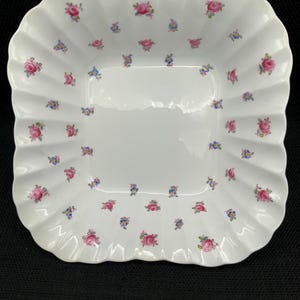 Könnte beinhalten: Weißer quadratischer Servierteller mit gewelltem Rand, verziert mit einem Streumuster aus rosa Rosen und kleinen blauen Blumen. Der Teller hat eine glänzende Oberfläche, ideal zum Servieren von Vorspeisen oder Desserts.