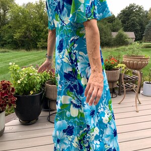 Puede incluir: Un vestido largo hasta el suelo con un vibrante estampado floral en tonos azul brillante, azul marino y verde lima. La prenda tiene mangas cortas acampanadas y un corpi&ntilde;o ajustado que se transforma en una falda fluida con dise&ntilde;o bot&aacute;nico.