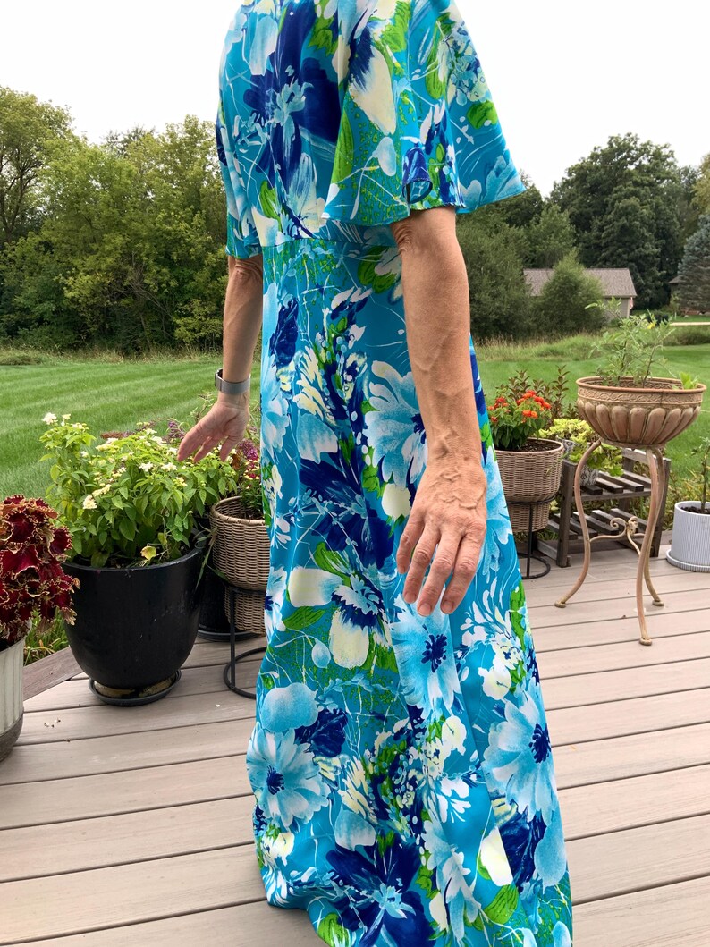 Puede incluir: Un vestido largo hasta el suelo con un vibrante estampado floral en tonos azul brillante, azul marino y verde lima. La prenda tiene mangas cortas acampanadas y un corpi&ntilde;o ajustado que se transforma en una falda fluida con dise&ntilde;o bot&aacute;nico.