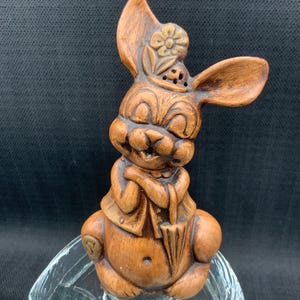 May include: A brown ceramic rabbit figurine with a flower on its head, holding an umbrella, sitting on a clear glass base.
