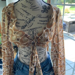 K&ouml;nnte beinhalten: Ein durchsichtiges, lang&auml;rmliges Crop-Top mit einem braunen und beigefarbenen geometrischen Muster. Das Oberteil ist an der Taille gebunden und wird mit Jeansshorts kombiniert. Das Oberteil wird auf einer Schaufensterpuppe pr&auml;sentiert.
