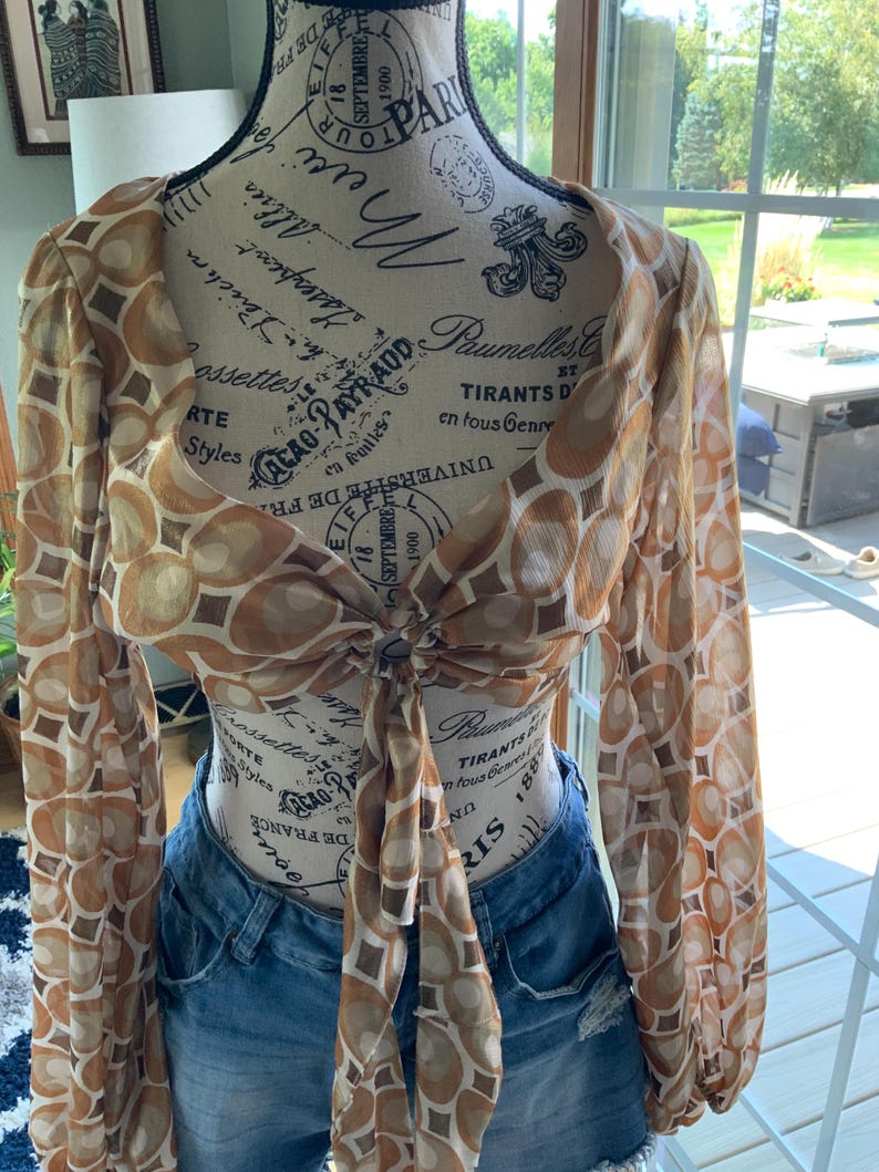 K&ouml;nnte beinhalten: Ein durchsichtiges, lang&auml;rmliges Crop-Top mit einem braunen und beigefarbenen geometrischen Muster. Das Oberteil ist an der Taille gebunden und wird mit Jeansshorts kombiniert. Das Oberteil wird auf einer Schaufensterpuppe pr&auml;sentiert.