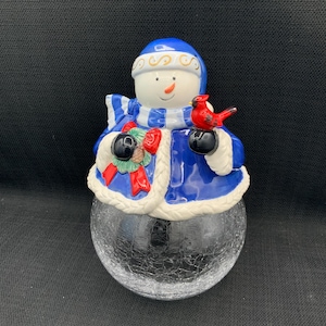 Op de afbeelding: Een keramieken sneeuwpopfiguur met een blauwe hoed en jas, met een rode kardinaal. De sneeuwpop heeft een wit gezicht met een wortelneus en zwarte handschoenen. De basis is een heldere, gekraakte glazen bol.