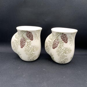 May include: Two off-white ceramic mugs featuring a repeating pattern of green pine needles and brown pinecones. Each mug has a rounded, protruding handle on the side and a smooth, curved body shape with a wide, flared rim at the top.
