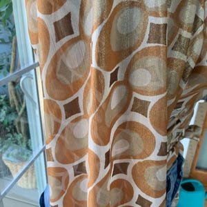 K&ouml;nnte beinhalten: Ein transparenter Stoff mit einem sich wiederholenden Muster aus braunen, goldenen und wei&szlig;en abstrakten Formen. Das Design umfasst gro&szlig;e Ovale und Rauten. Der Stoff ist drapiert, was darauf hindeutet, dass es sich um einen Schal, Vorhang oder ein Kleidungsst&uuml;ck handeln k&ouml;nnte.