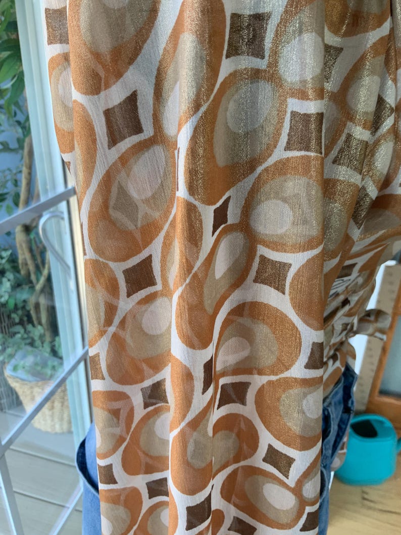 K&ouml;nnte beinhalten: Ein transparenter Stoff mit einem sich wiederholenden Muster aus braunen, goldenen und wei&szlig;en abstrakten Formen. Das Design umfasst gro&szlig;e Ovale und Rauten. Der Stoff ist drapiert, was darauf hindeutet, dass es sich um einen Schal, Vorhang oder ein Kleidungsst&uuml;ck handeln k&ouml;nnte.