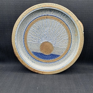 Puede incluir: Un plato redondo de cerámica con un diseño de sol. El plato presenta una superficie texturizada y moteada con una combinación de colores azul claro y marrón. El diseño del sol está en el centro, con un borde azul y marrón.