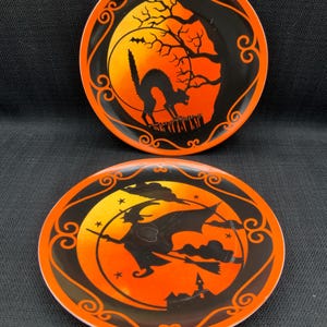 May include: Two Halloween-themed decorative plates. One plate features a black cat silhouette against an orange and yellow background with a tree and a bat. The other plate shows a witch on a broomstick, a castle, and stars.