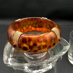 May include: A thick, translucent amber-colored bangle bracelet featuring a repeating dark brown spotted pattern. The bracelet is accented with two gold-toned metal bands that have a textured mesh surface and small, round, dark amber-colored studs.