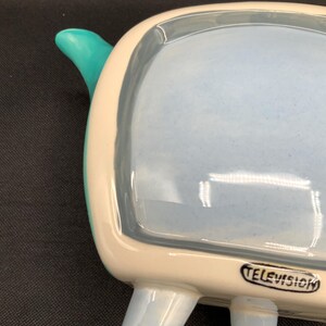May include: A ceramic television-shaped dish with a light blue interior and legs. The TV has a white border and a teal handle. The word "TELEVISION" is printed on the front. A retro-style decorative piece.