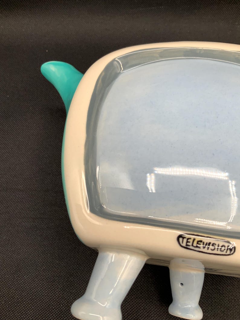 May include: A ceramic television-shaped dish with a light blue interior and legs. The TV has a white border and a teal handle. The word "TELEVISION" is printed on the front. A retro-style decorative piece.