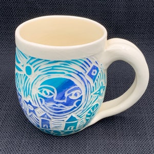 Könnte beinhalten: Weiße Keramiktasse mit einem blauen und grünen Design. Das Design zeigt ein stilisiertes Gesicht mit Augen und Mund, umgeben von Wirbeln und Linien. Es gibt auch kleine Häuser und Sterne im Design.