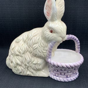 May include: A white ceramic bunny figurine with pink ears and red eyes, holding a purple and white basket.