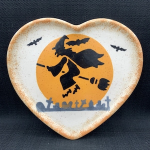 Peut inclure: Assiette en céramique en forme de cœur sur le thème d'Halloween. L'assiette présente une silhouette noire d'une sorcière volant sur un balai devant une lune orange. Comprend des chauves-souris et une scène de cimetière. Fond blanc moucheté et bord marron.
