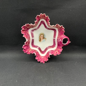 May include: A decorative, antique porcelain dish with a star-shaped design. The dish features a white interior with a portrait of a woman, framed by a magenta border and gold accents. The outer edge has a ruffled, scalloped design.