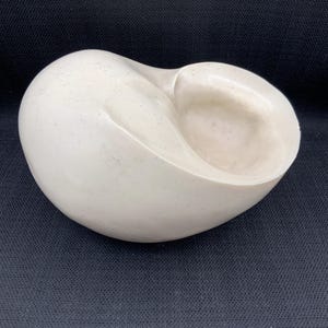Può includere: Scultura liscia bianco sporco dalla forma arrotondata e organica. Presenta una profonda rientranza curva su un lato, creando una forma a ciotola. La superficie presenta sottili variazioni di colore e consistenza.