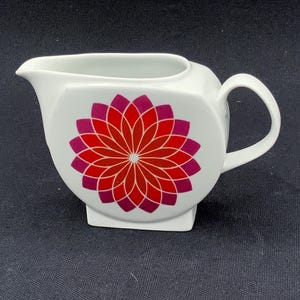 Puede incluir: Una jarra de leche de cerámica blanca con un patrón floral geométrico en tonos rojo y magenta en el lateral. La jarra cuenta con un asa curva, un pico vertedor puntiagudo y una base plana, mostrando un diseño limpio y funcional.