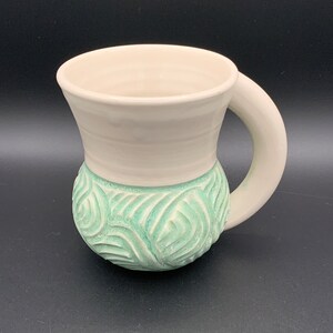 May include: A white ceramic mug with a green, leafy design. The mug has a rounded body and a curved handle.