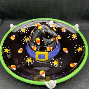 Puede incluir: Un plato de cerámica negro para servir en Halloween con un diseño de sombrero de bruja. El sombrero está decorado con caramelos de maíz y un cinturón azul. El plato tiene un borde verde, estrellas blancas y pequeños fantasmas.