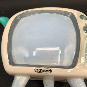 May include: A ceramic television-shaped dish with a light blue interior and a white and light blue exterior. The word "TELEVISION" is printed on the bottom. The dish has four light blue legs and a teal handle.