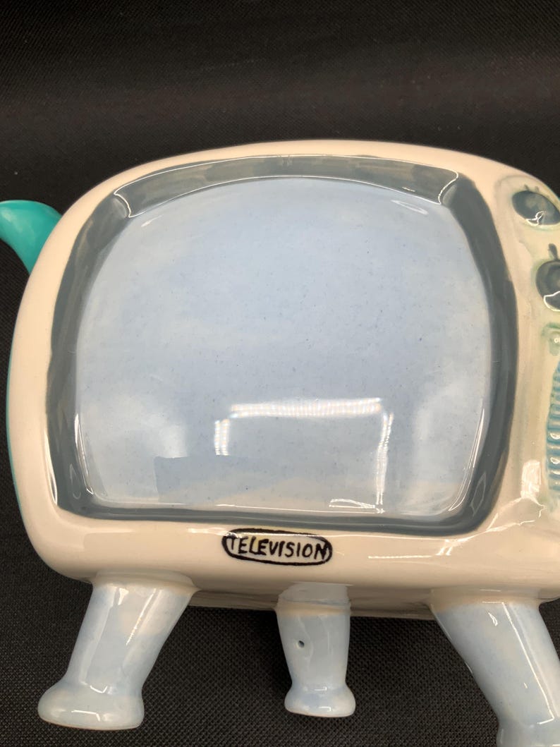 May include: A ceramic television-shaped dish with a light blue interior and a white and light blue exterior. The word "TELEVISION" is printed on the bottom. The dish has four light blue legs and a teal handle.