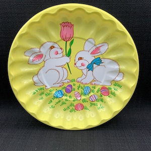 May include: A yellow ceramic plate with a scalloped edge features two white bunnies holding a pink tulip. The bunnies are surrounded by green grass and colorful Easter eggs.
