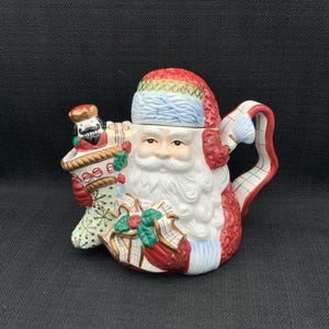 Puede incluir: Una tetera de cerámica de Papá Noel con gorro y barba rojos, sosteniendo un cascanueces. La tetera presenta un diseño a cuadros rojos y blancos en la base y el asa, con detalles en verde y dorado. Una decoración festiva para las fiestas.