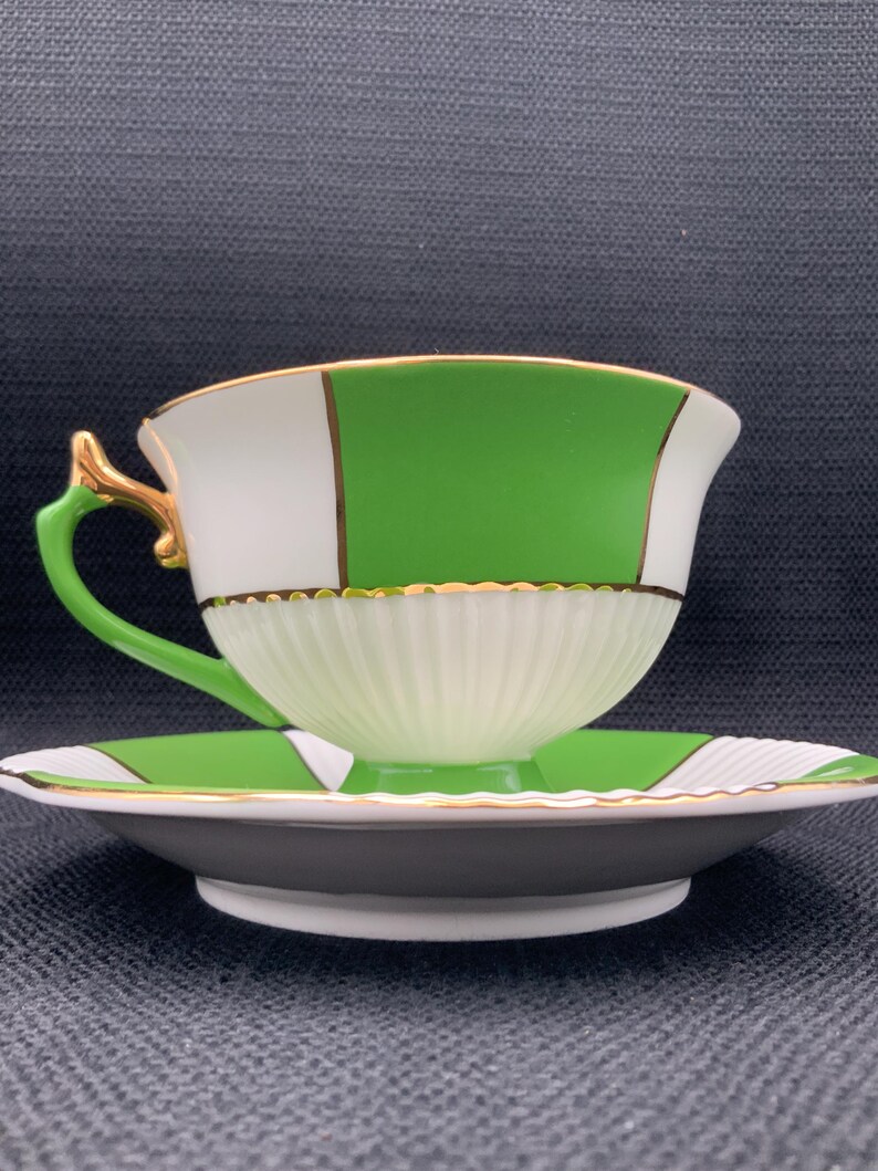 May include: A white and green teacup and saucer set with gold trim. The cup and saucer have a geometric pattern with white, green, and gold accents.