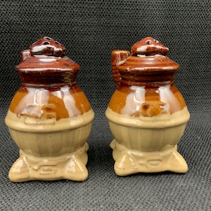 May include: Two ceramic salt and pepper shakers with a brown and tan glaze. Each shaker has a rounded body, a small handle, and a lid with holes. The shakers are on a dark background.