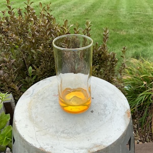 May include: A clear glass tumbler with a thick, rounded base featuring a vibrant amber-colored tint. The glass has straight, smooth sides and a wide, open rim, designed for holding beverages, showcasing a simple and functional cylindrical shape.