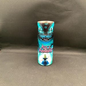 May include: A cylindrical tumbler featuring a vibrant design from "Alice in Wonderland." The design includes the Cheshire Cat's grin and the words "Alice Wonderland." The tumbler is predominantly blue and black.