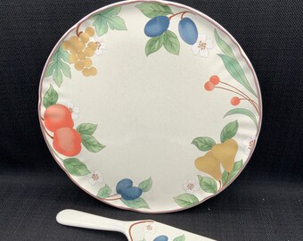 May include: A white ceramic cake plate with a fruit design. The plate has a scalloped edge and features a border of grapes, pears, apples, and plums. The plate comes with a matching white ceramic cake server.