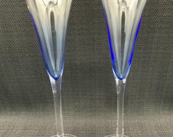 Puede incluir: Dos copas de champán transparentes con un acento azul en el interior del cristal. Las copas tienen un tallo largo y delgado y una base redonda. Las copas son de cristal y están diseñadas para servir champán o vino espumoso.