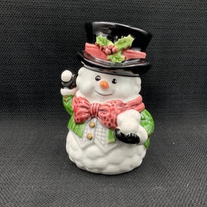 May include: A ceramic snowman figurine wearing a black top hat with holly, a red bow tie, and a green vest. The snowman has a carrot nose, black eyes, and a smiling mouth. The figurine is a festive holiday decoration.