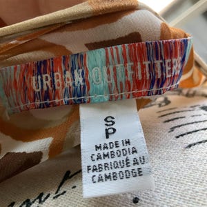 K&ouml;nnte beinhalten: Nahaufnahme eines Etiketts von Urban Outfitters. Das wei&szlig;e Etikett tr&auml;gt den schwarzen Text "S", "Made in Cambodia" und "Fabriqu&eacute; au Cambodge". Der Stoff hat ein braunes und wei&szlig;es Muster.
